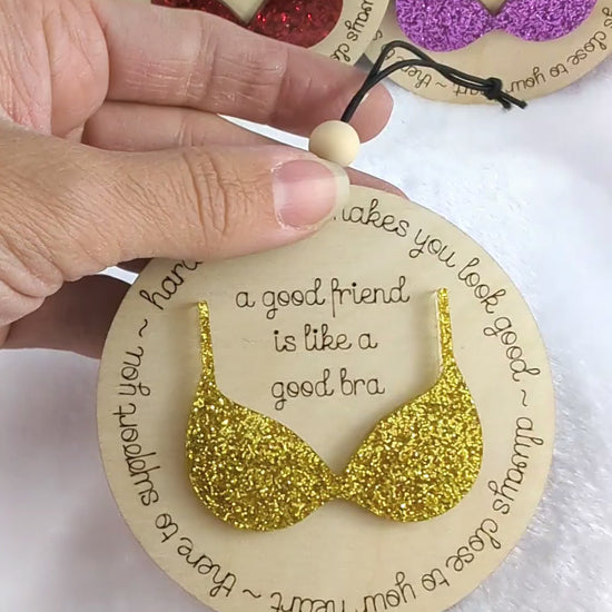 Video of Friendship ornament with a bra. Reads: A good friend is like a good bra. Shows 4 different color options. Red, purple, green and gold glitter bras. 