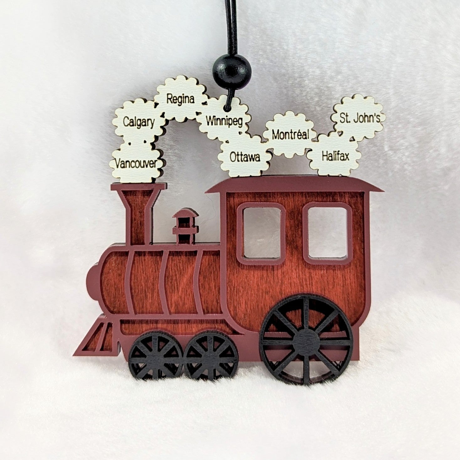A personalized wooden Christmas train ornament with a reddish-brown wood grain finish, black wheels, and white cloud-shaped steam details printed with city names including Calgary, Vancouver, Winnipeg, Ottawa, Montreal, St. John's, and Halifax.