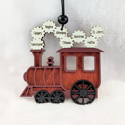 A personalized wooden Christmas train ornament with a reddish-brown wood grain finish, black wheels, and white cloud-shaped steam details printed with city names including Calgary, Vancouver, Winnipeg, Ottawa, Montreal, St. John's, and Halifax.