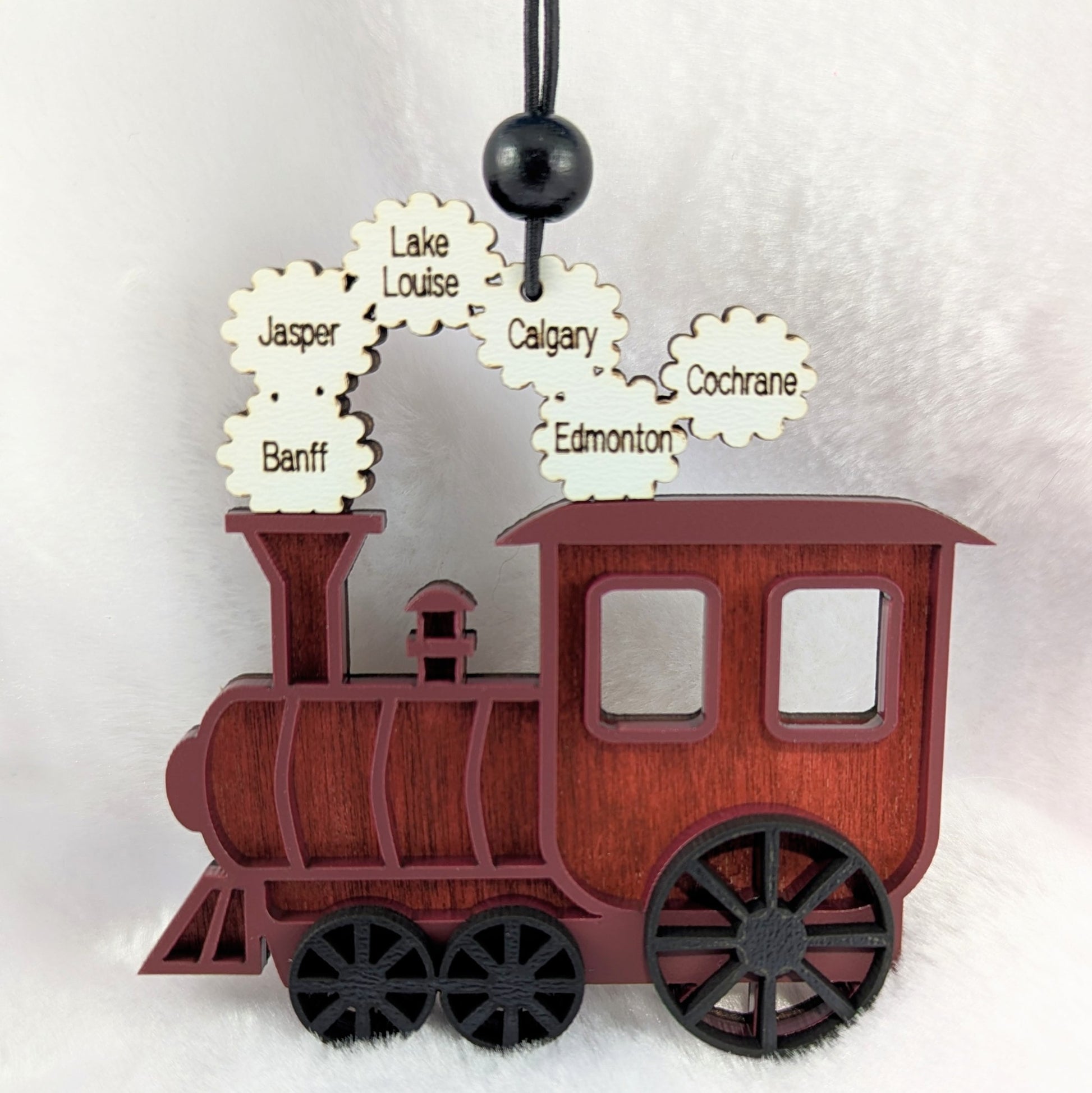 A personalized wooden train ornament with a dark red-brown body, black wheels and hanging bead, and cream-colored smokestack cutouts engraved with names like Jasper, Banff, Calgary, Edmonton, Lake Louise, and Cochrane, shown against a soft white background.