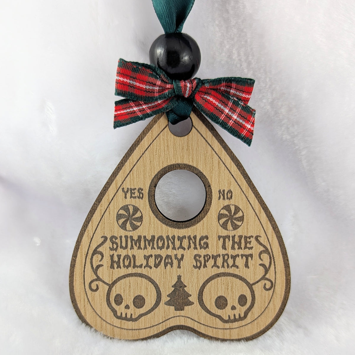 A wooden planchette-shaped ornament with engraved text reading 'SUMMONING THE HOLIDAY SPIRIT' and skull, candy cane, and Christmas tree illustrations. It has a red and green plaid ribbon bow with a dark green hanging ribbon and black beads at the top, shown against a white textured background.